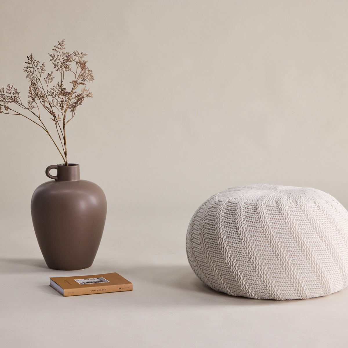 Ted puff rund i offwhite strikk, ved brun vase med tørrblomster og bok