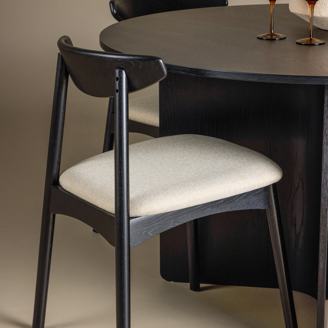Ven spisestol i sort eik med beige sete ved mørkt rundt bord med ravfargede glass.