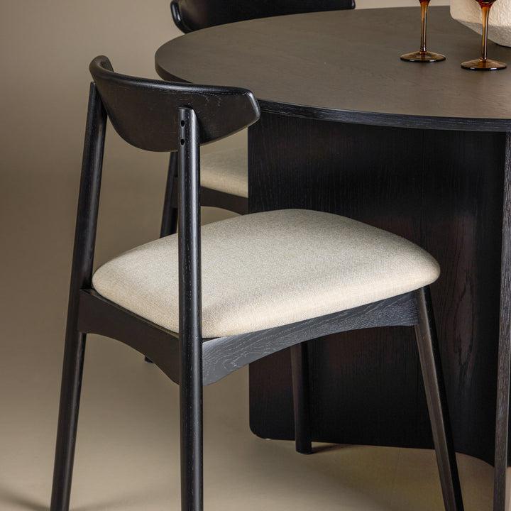Ven spisestol i sort eik med beige sete ved mørkt rundt bord med ravfargede glass.