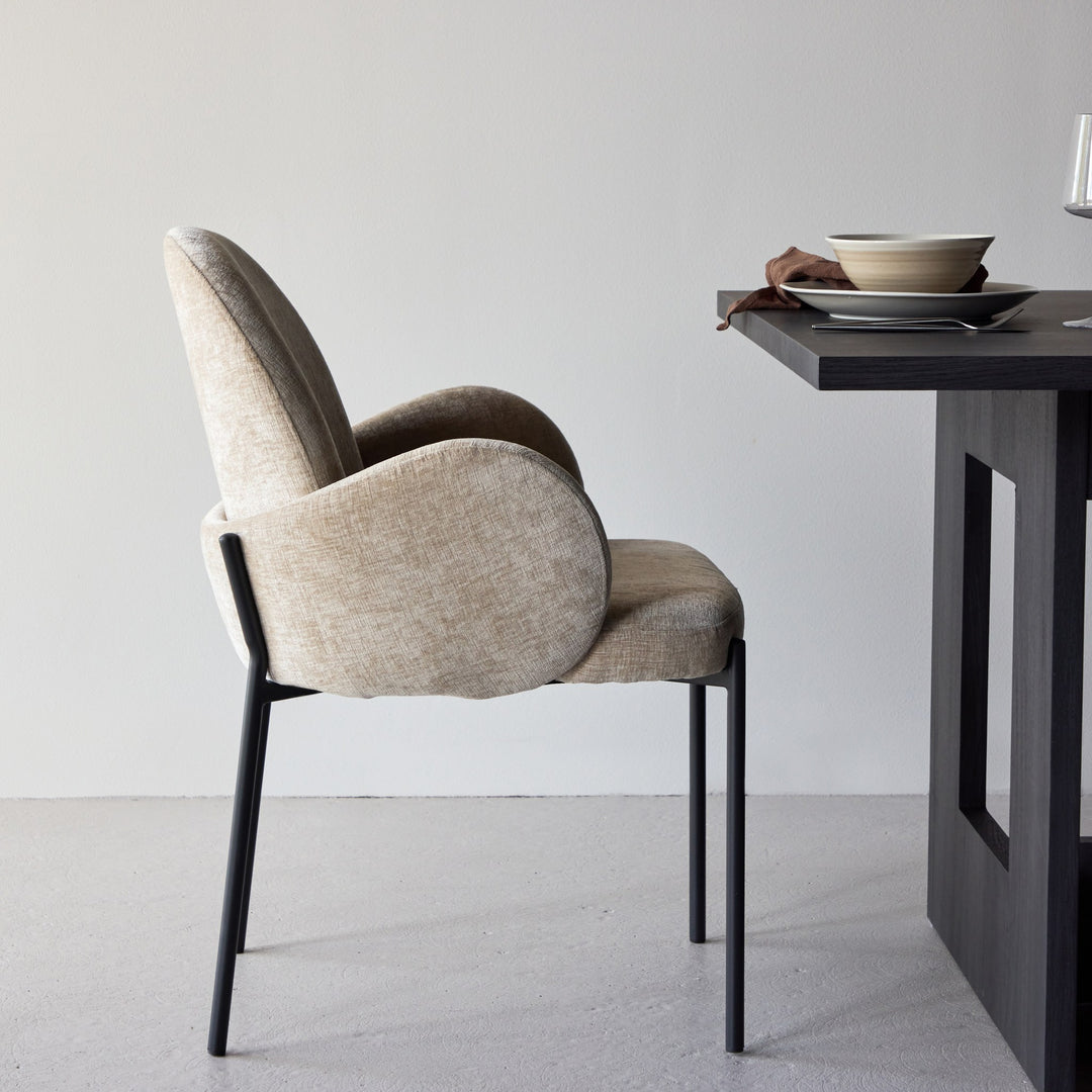 Blom spisestol i beige chenille med sorte metallben, sett fra siden ved mørkt spisebord.