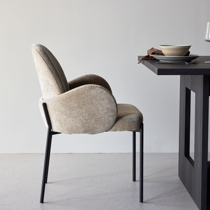 Blom spisestol i beige chenille med sorte metallben, sett fra siden ved mørkt spisebord.