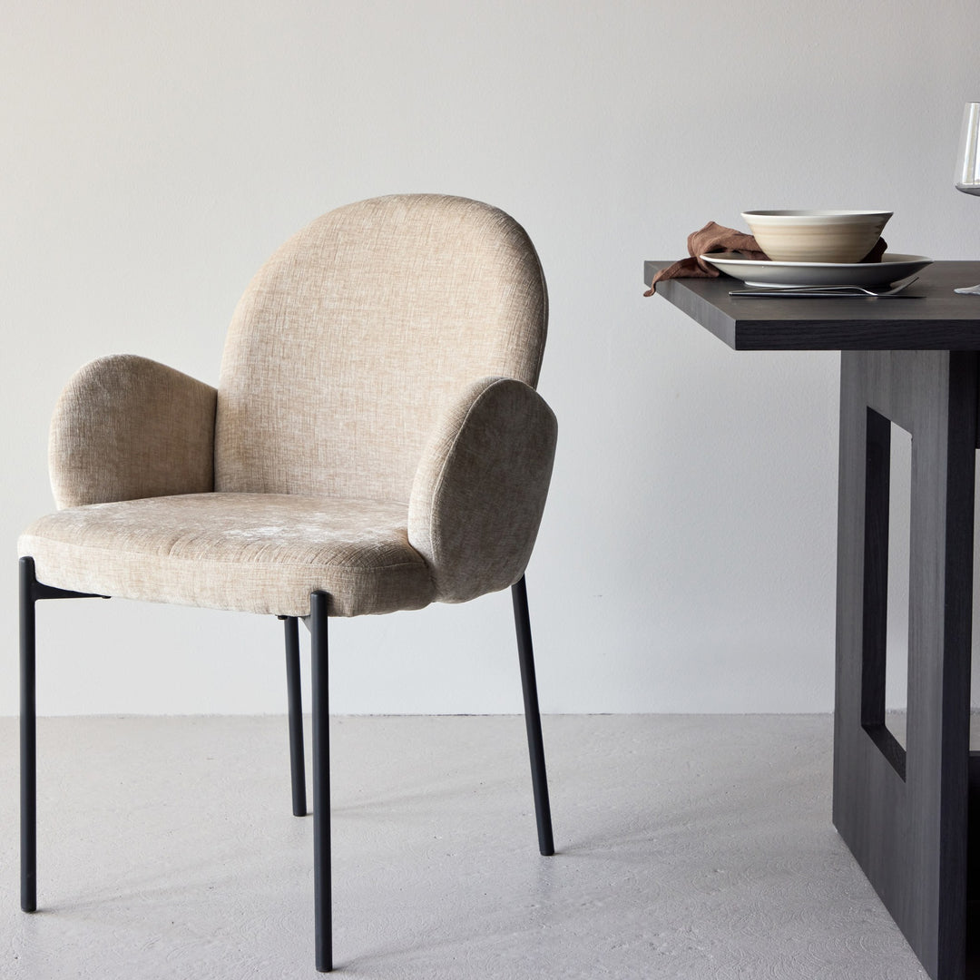 Blom spisestol i beige chenille med buet rygg og armlener, sorte ben, ved mørkt bord