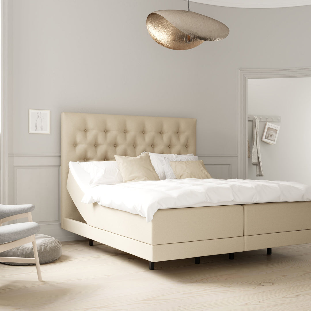 Wonderland Pique sengegavl i lys beige stoff med knappet design, bak oppredd seng med gull taklampe.