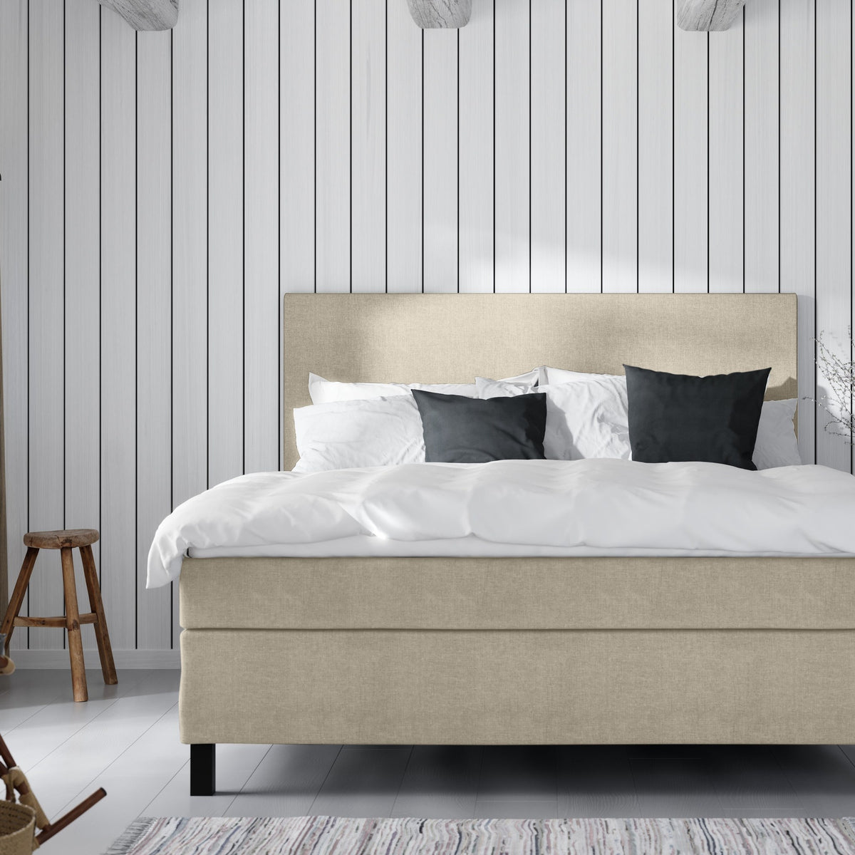 Wonderland W50 sengegavl i beige stoff, hvit seng med mørkegrå puter mot hvit panelvegg, trekrakk og fillerye.