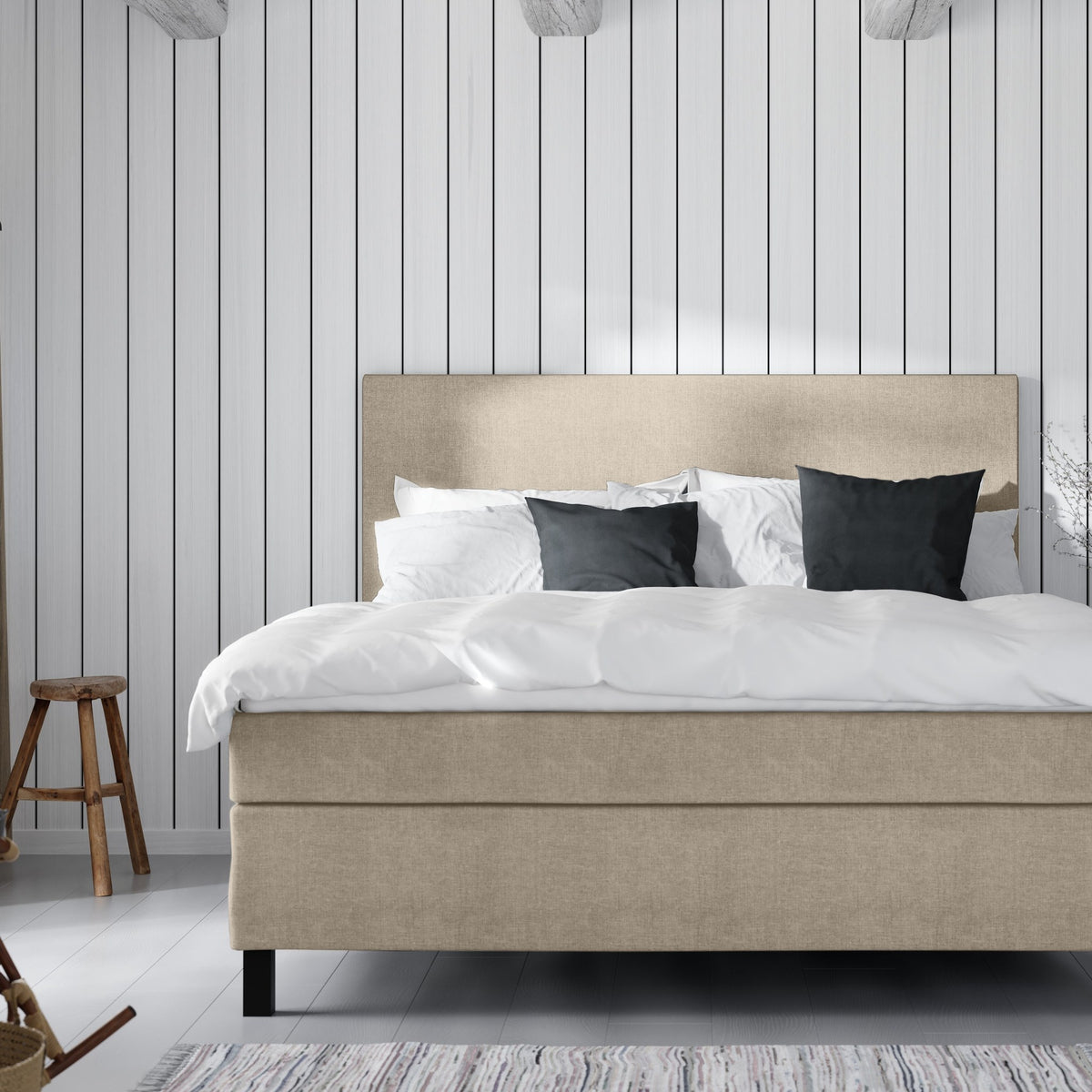 Wonderland W50 sengegavl i sandbeige stoff, hvit seng med mørke puter mot hvit panelvegg og trekrakk.
