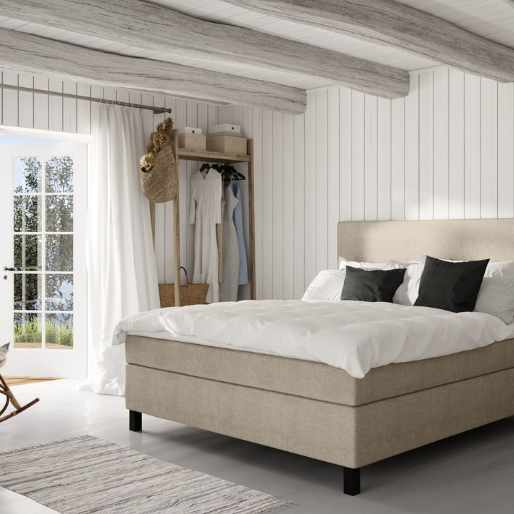 Wonderland W50 sengegavl i gråbeige, oppredd seng i lyst panelt soverom med klesstativ og teppe
