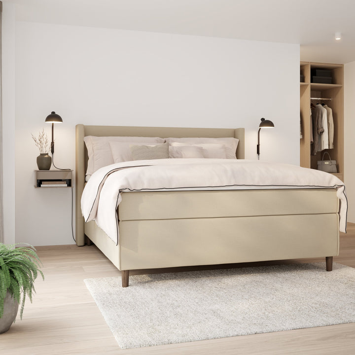 Wonderland Wing sengegavl i lys beige stoff, oppredd seng med vegglamper, nattbord og grønn plante i soverom.