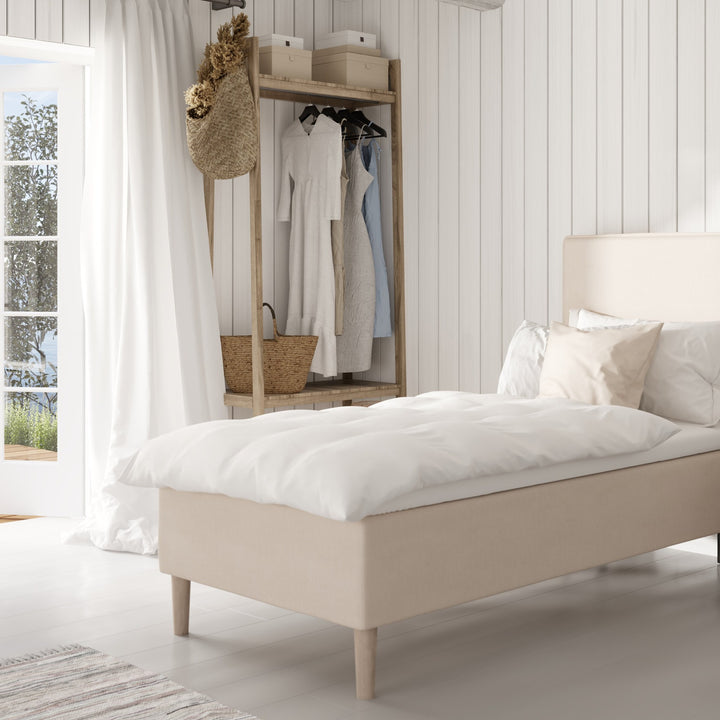 Wonderland Lento rammeseng i sandbeige stoff med hvit dyne, mot hvit panelvegg med åpen garderobe og vindu.