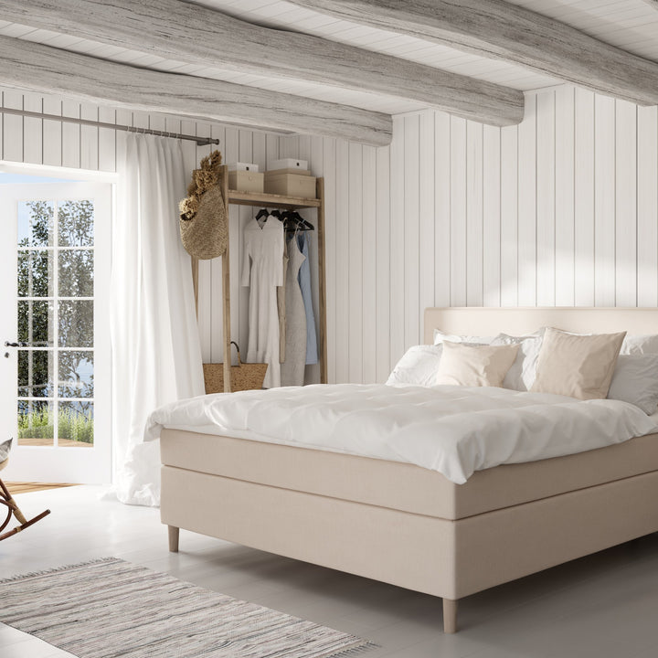 Wonderland Note kontinentalseng i sandbeige stoff med hvit dyne, i lyst panelrom med åpen garderobe.