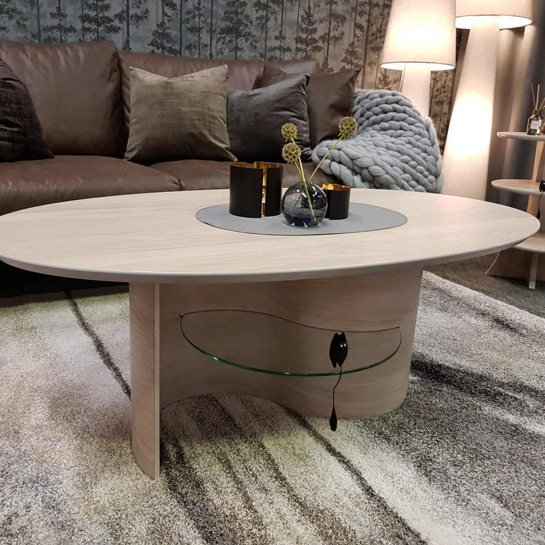 Esset sofabord i lyst tre, ovalt med glasshylle, i stue med brun skinnsofa og gulvlampe